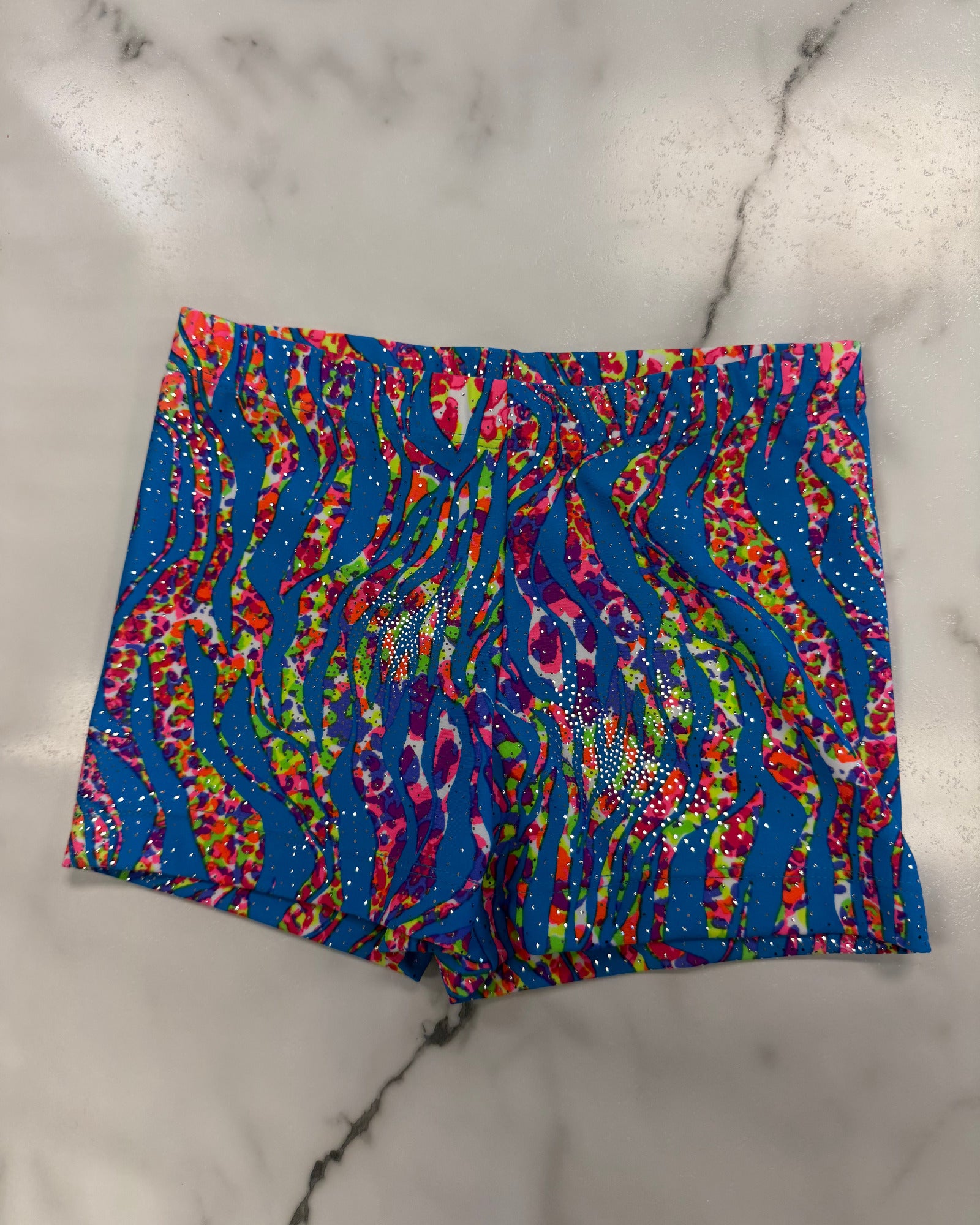 A pair of women's dance shorts featuring a vibrant, multicolored abstract pattern with blue, pink, red, green, and yellow swirls, laid flat on a white marble surface.