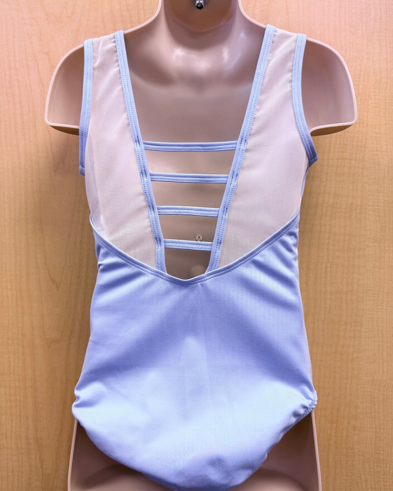 A light blue and white sleeveless leotard displayed on a beige mannequin torso against a light wood background.