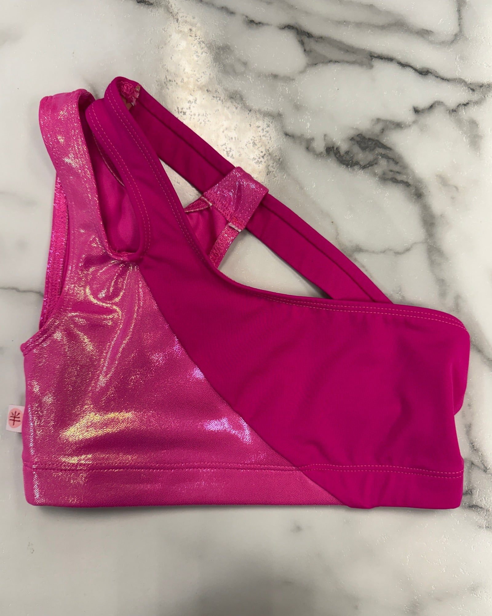 A bright pink dance bra with a shiny metallic magenta panel and solid pink side panels, laid flat on a white marble surface.