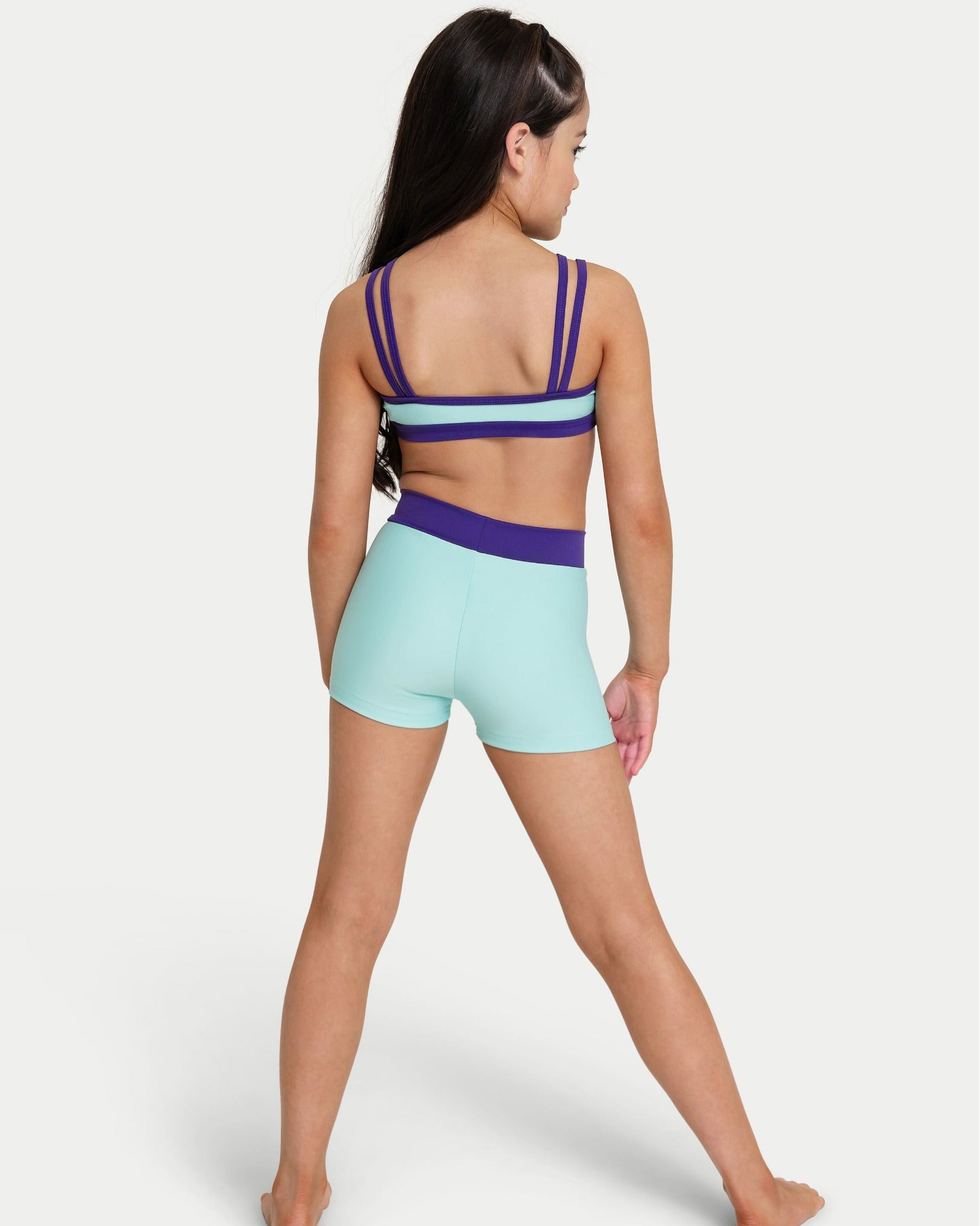 A girl modeling a two-piece activewear set from the back, featuring a mint green  bra top with blue straps and matching high-waisted mint green shorts with a purple waistband, set against a plain light gray background.