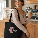 Woman holding a coffee cup and a black tote bag with 'Dead Pointe Shoe Club' text in a cafe.