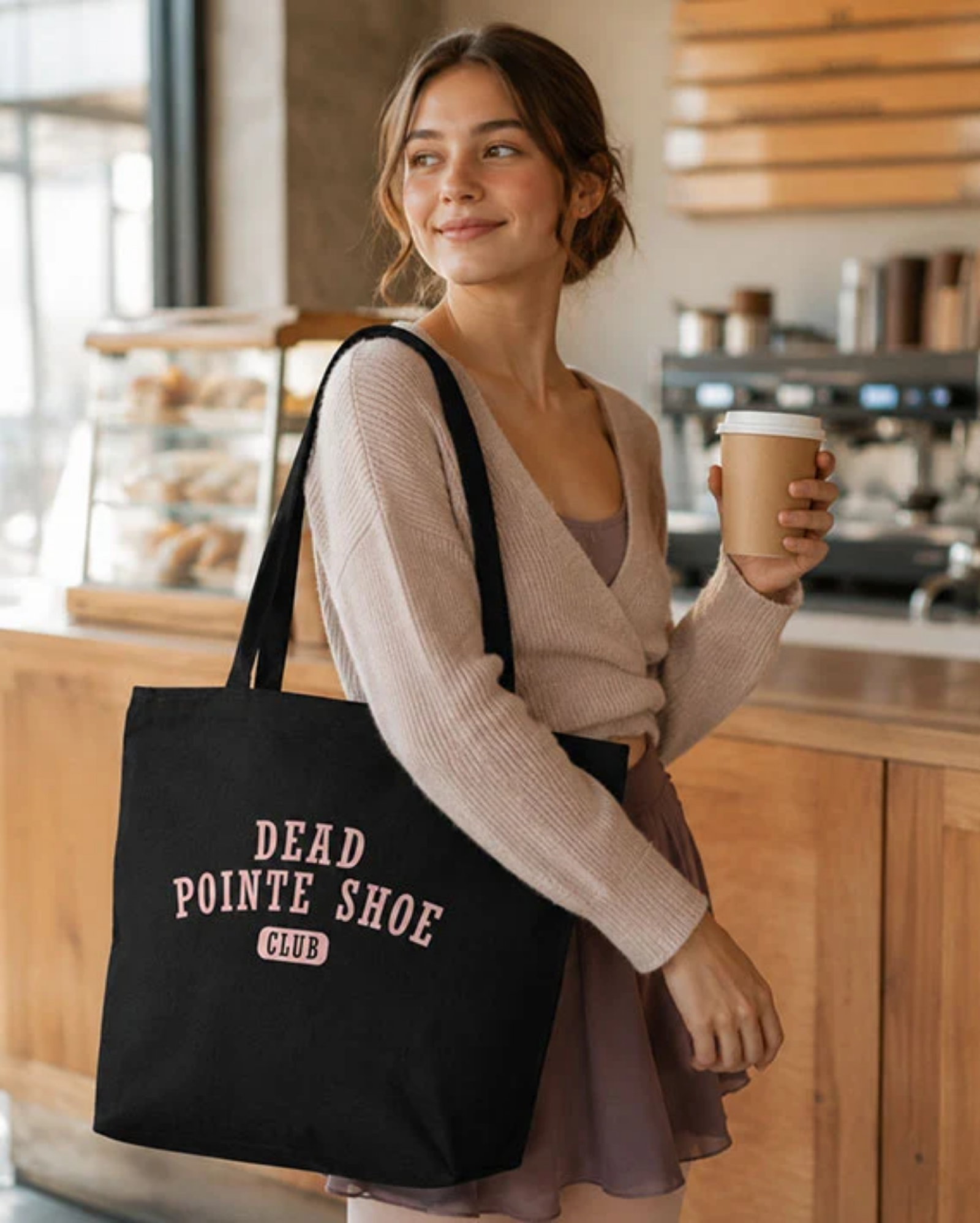 Woman holding a coffee cup and a black tote bag with 'Dead Pointe Shoe Club' text in a cafe.