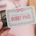 Hand holding a small tin labeled 'emergency bobby pins' with decorative design.