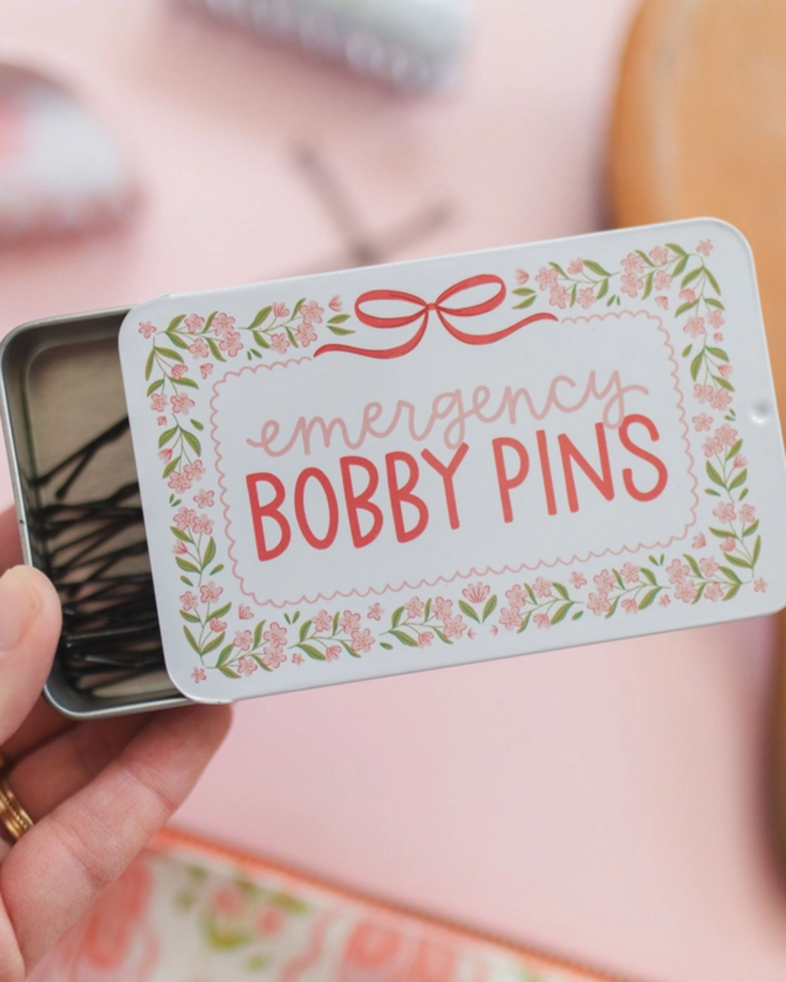 Hand holding a small tin labeled 'emergency bobby pins' with decorative design.