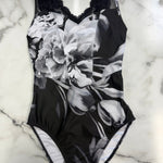 A black and white floral print one-piece leotard with black lace trim at the neckline and leg openings, laid flat on a white marble surface.