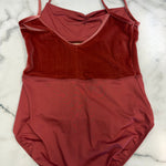 A rust-red two-tone leotard laid flat on a white marble surface. The top section is a textured, velvety rust red, while the bottom section is a smoother, matte brick red.