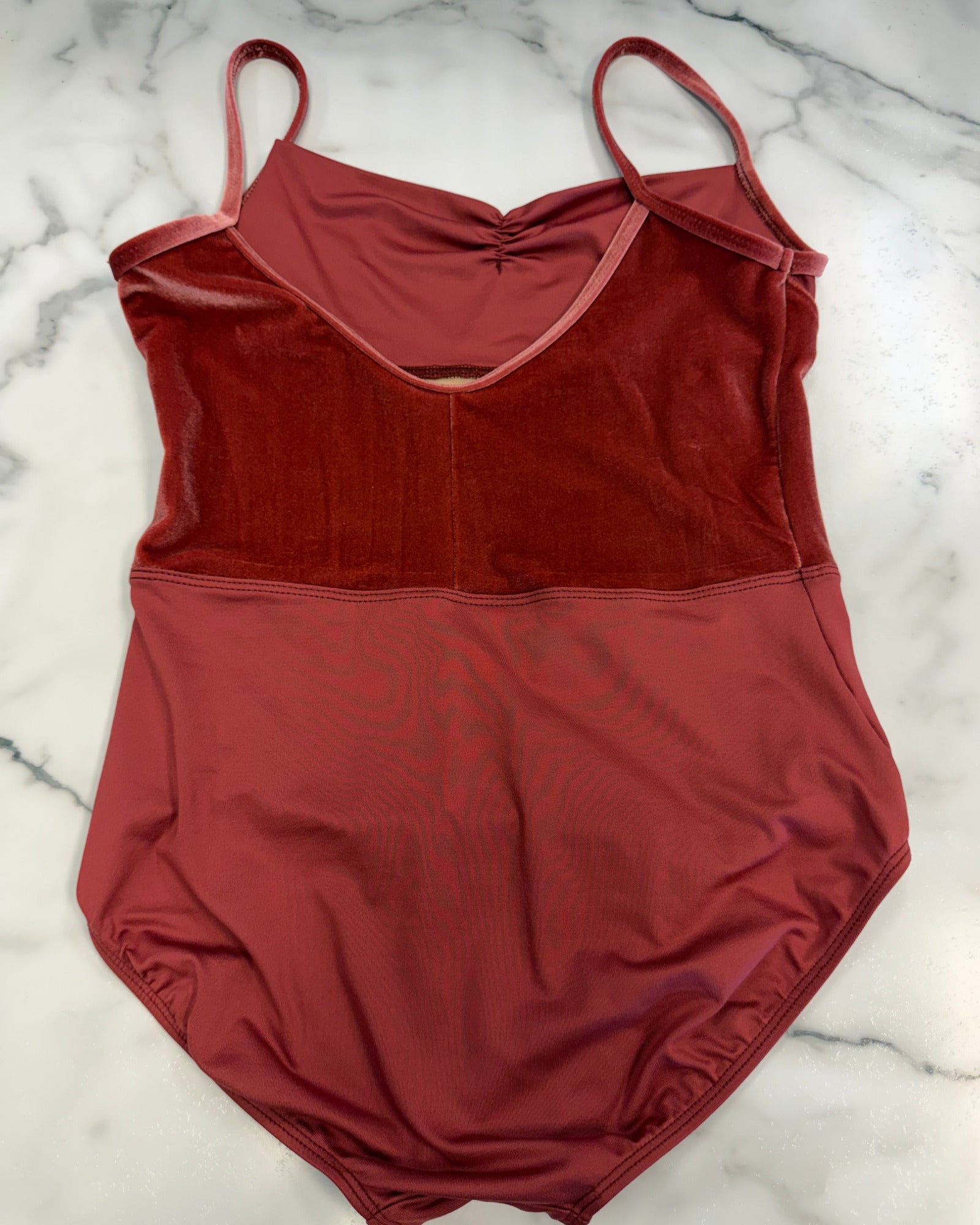 A rust-red two-tone leotard laid flat on a white marble surface. The top section is a textured, velvety rust red, while the bottom section is a smoother, matte brick red.