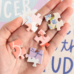 A hand holding five small pastel jigsaw puzzle pieces with floral and abstract designs, set against a softly blurred puzzle background.