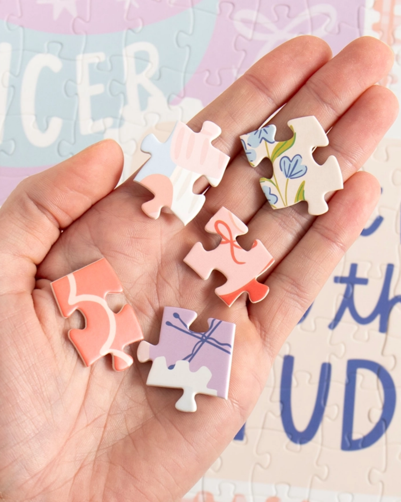 A hand holding five small pastel jigsaw puzzle pieces with floral and abstract designs, set against a softly blurred puzzle background.