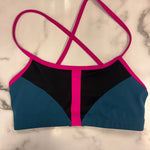 A color-blocked dance bra top laid flat on a white marble surface. The dance bra features bright pink straps and trim, a black center panel, and teal blue side panels.