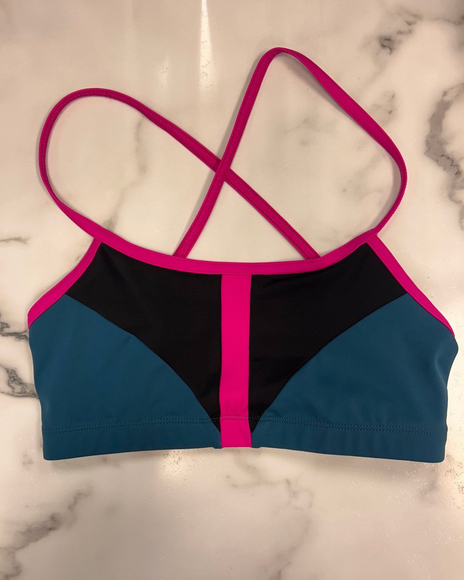 A color-blocked dance bra top laid flat on a white marble surface. The dance bra features bright pink straps and trim, a black center panel, and teal blue side panels.