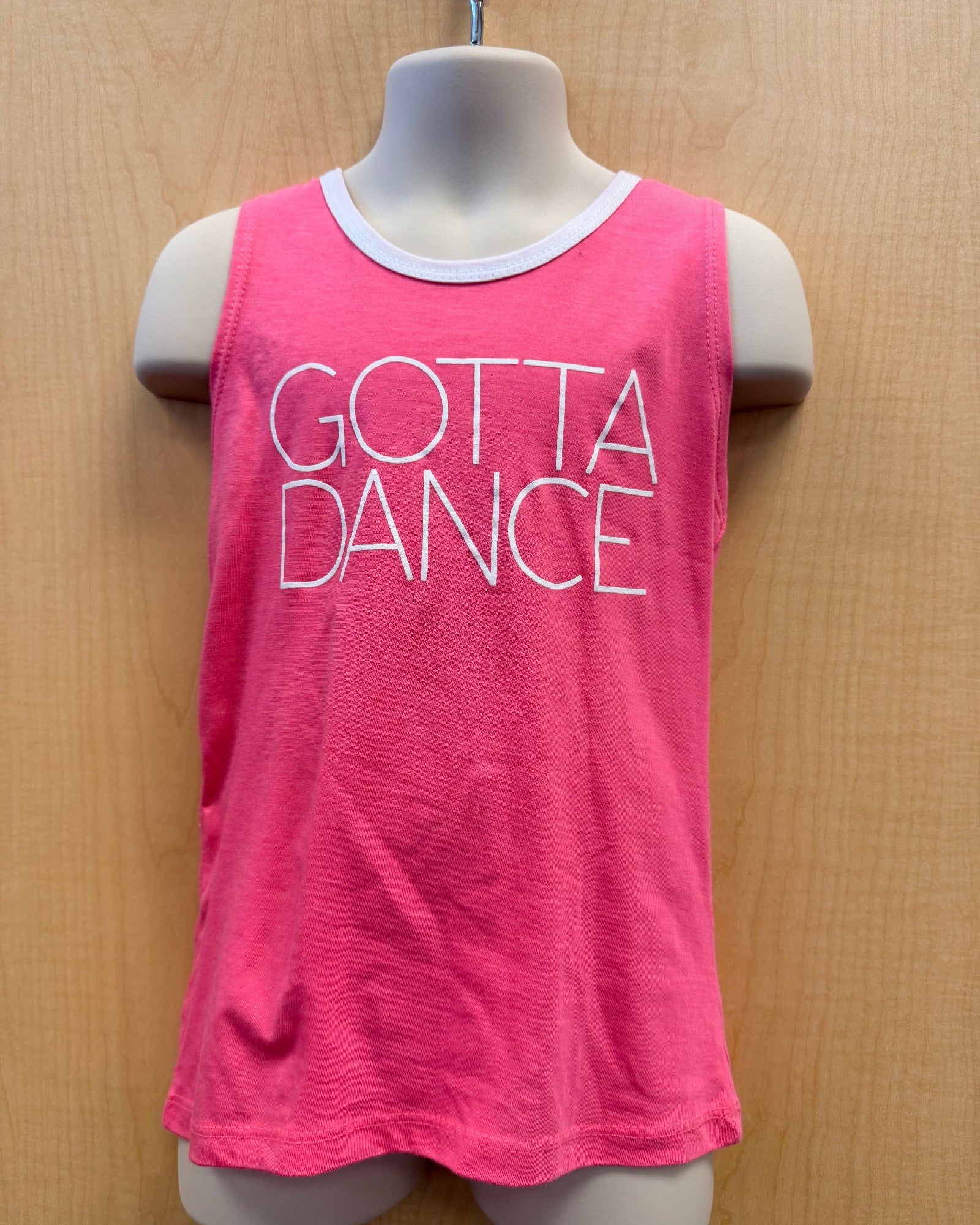 A bright pink sleeveless tank top with 'GOTTA DANCE' printed in white outline text across the front, displayed on a mannequin torso against a light wood background.