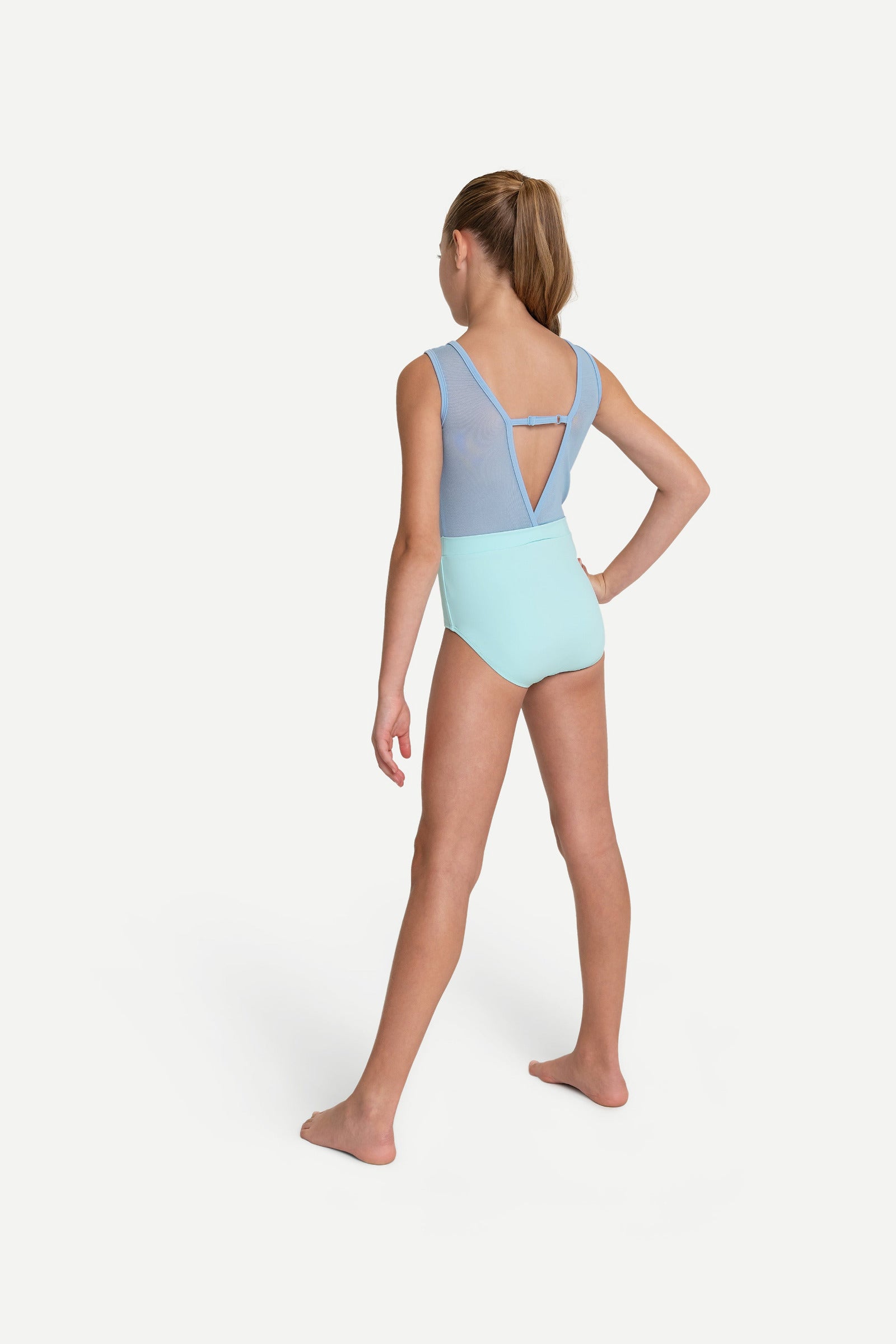 A young girl modeling a two-tone leotard from the back, featuring a light blue upper back and shoulder straps with a light mint green lower body, set against a plain light gray studio background.