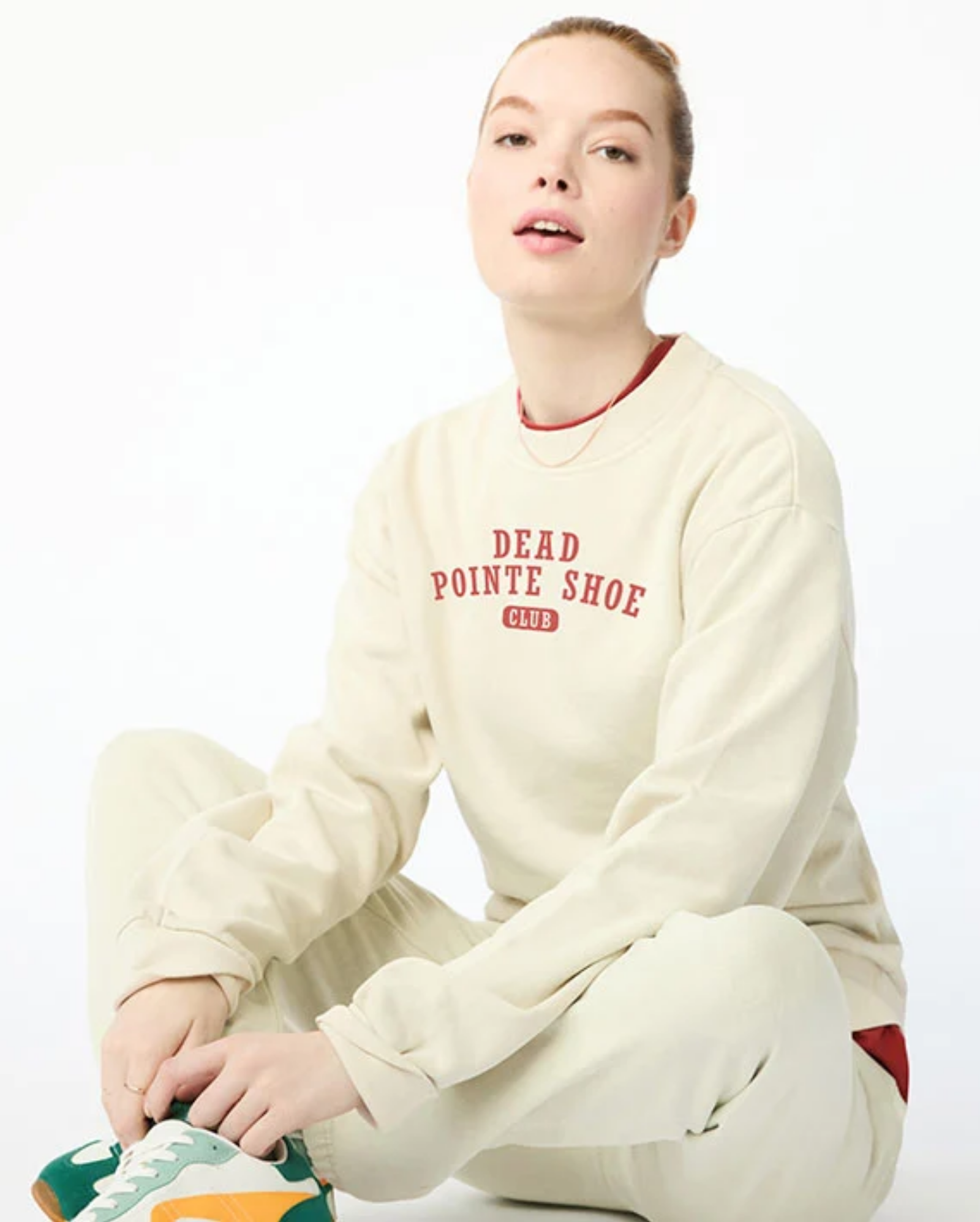 Person wearing a cream sweatshirt with text and pants, sitting on a white background