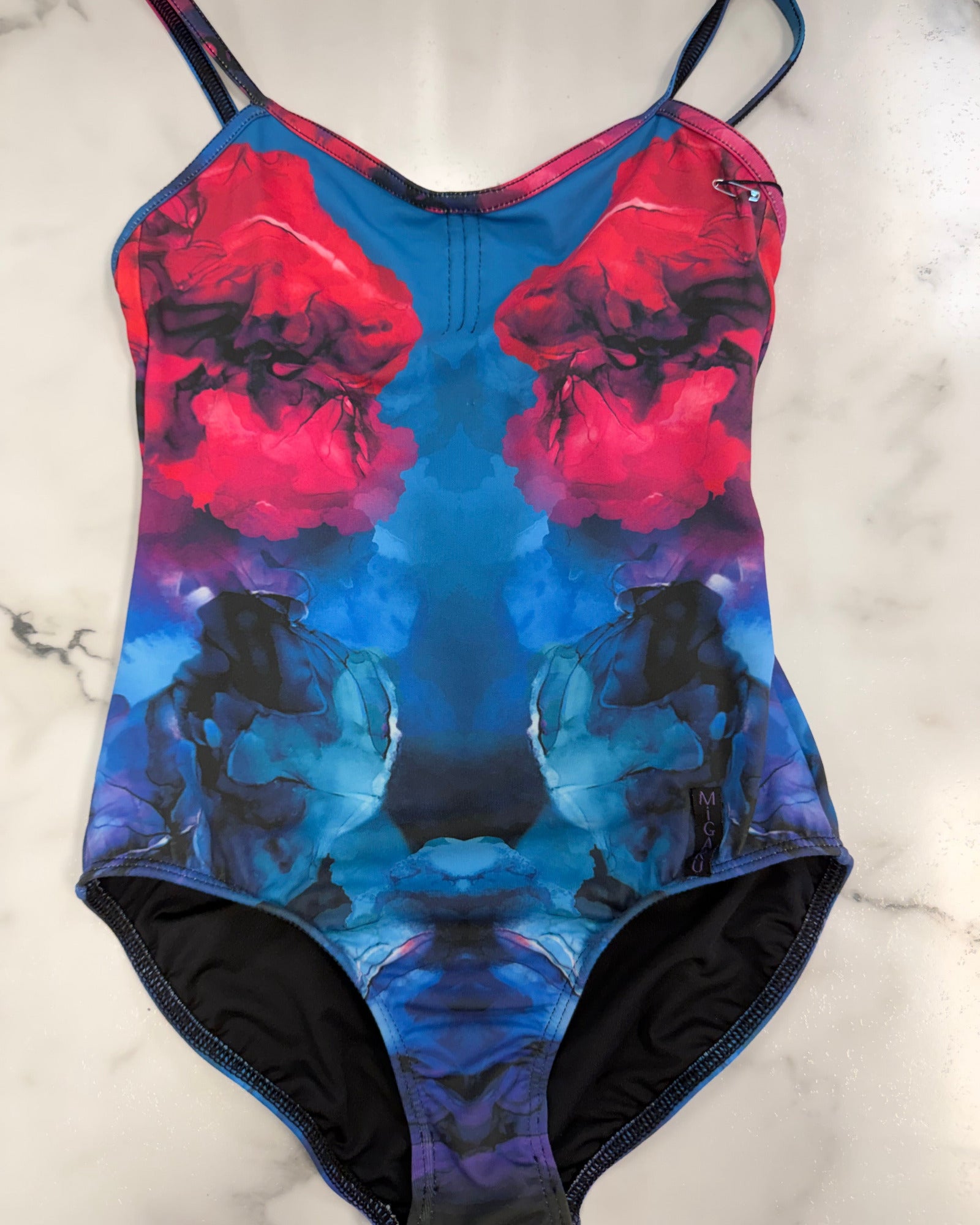 A leotard with a vibrant abstract print featuring red, pink, blue, and black watercolor-like shapes, laid flat on a white marble surface.