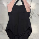 A colorblock leotard laid flat on a white marble background. The leotard is black on the lower body and upper chest, with light pink panels on the upper chest and shoulders, and black crisscross halter neck straps.