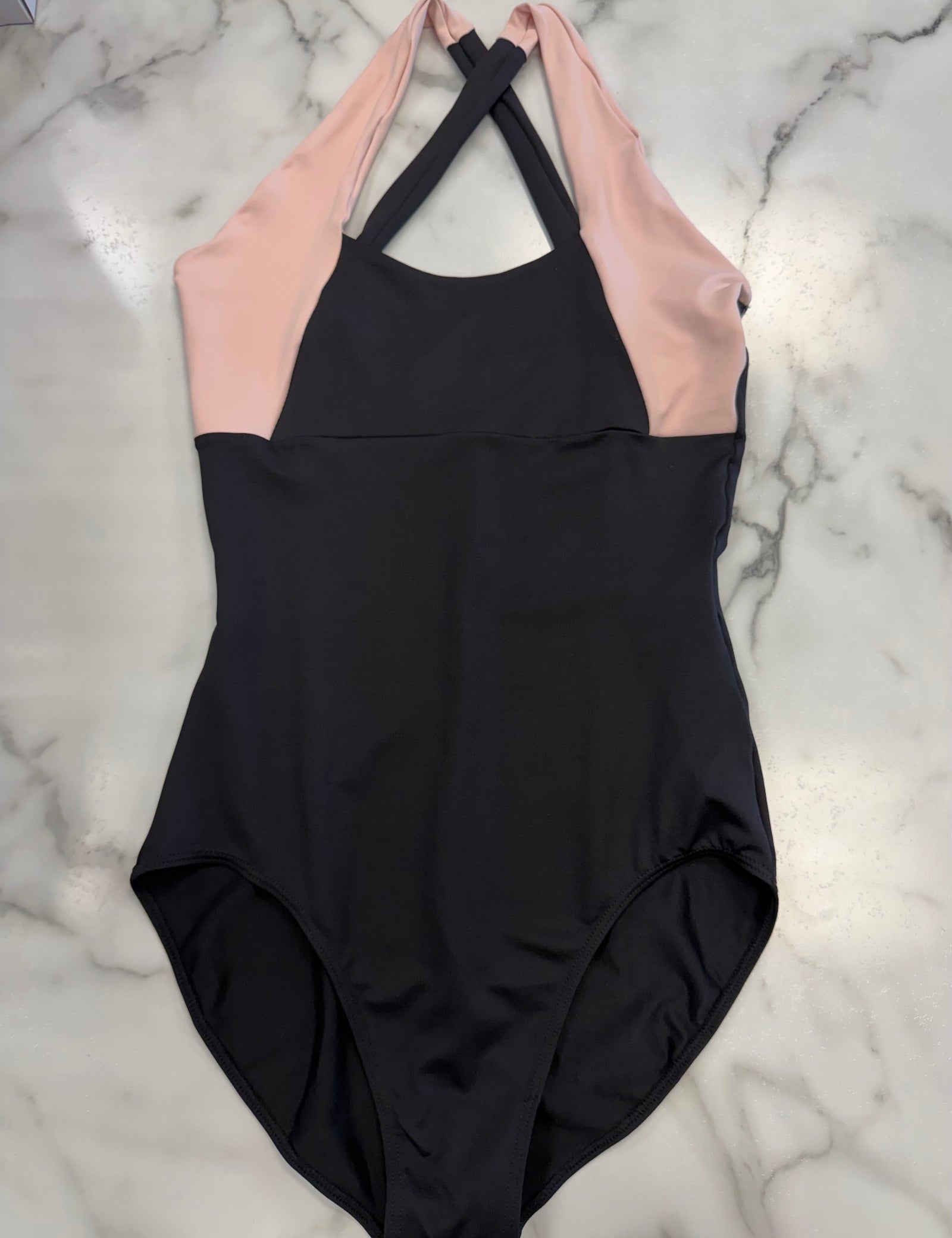 A colorblock leotard laid flat on a white marble background. The leotard is black on the lower body and upper chest, with light pink panels on the upper chest and shoulders, and black crisscross halter neck straps.