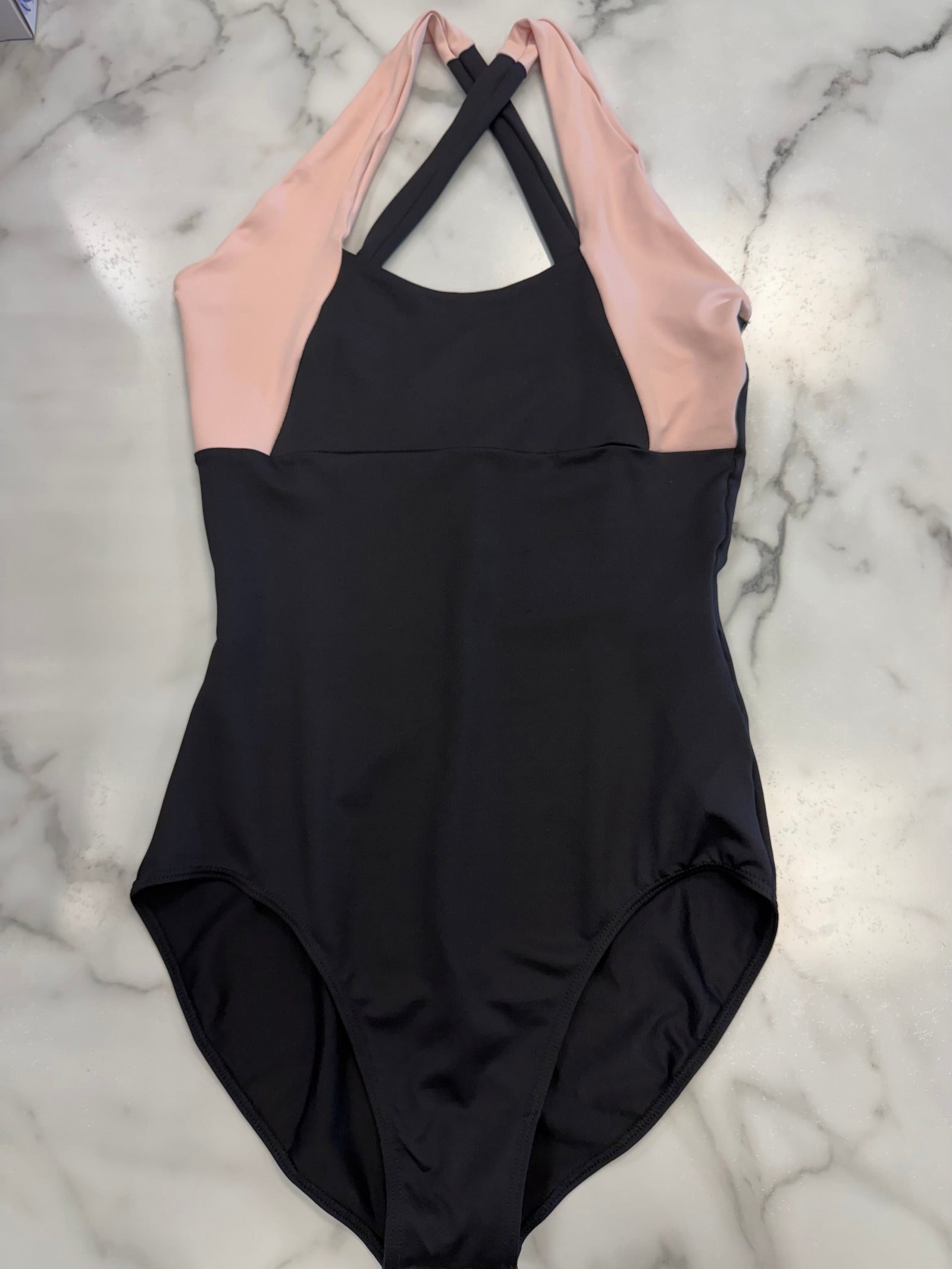 A colorblock leotard laid flat on a white marble background. The leotard is black on the lower body and upper chest, with light pink panels on the upper chest and shoulders, and black crisscross halter neck straps.