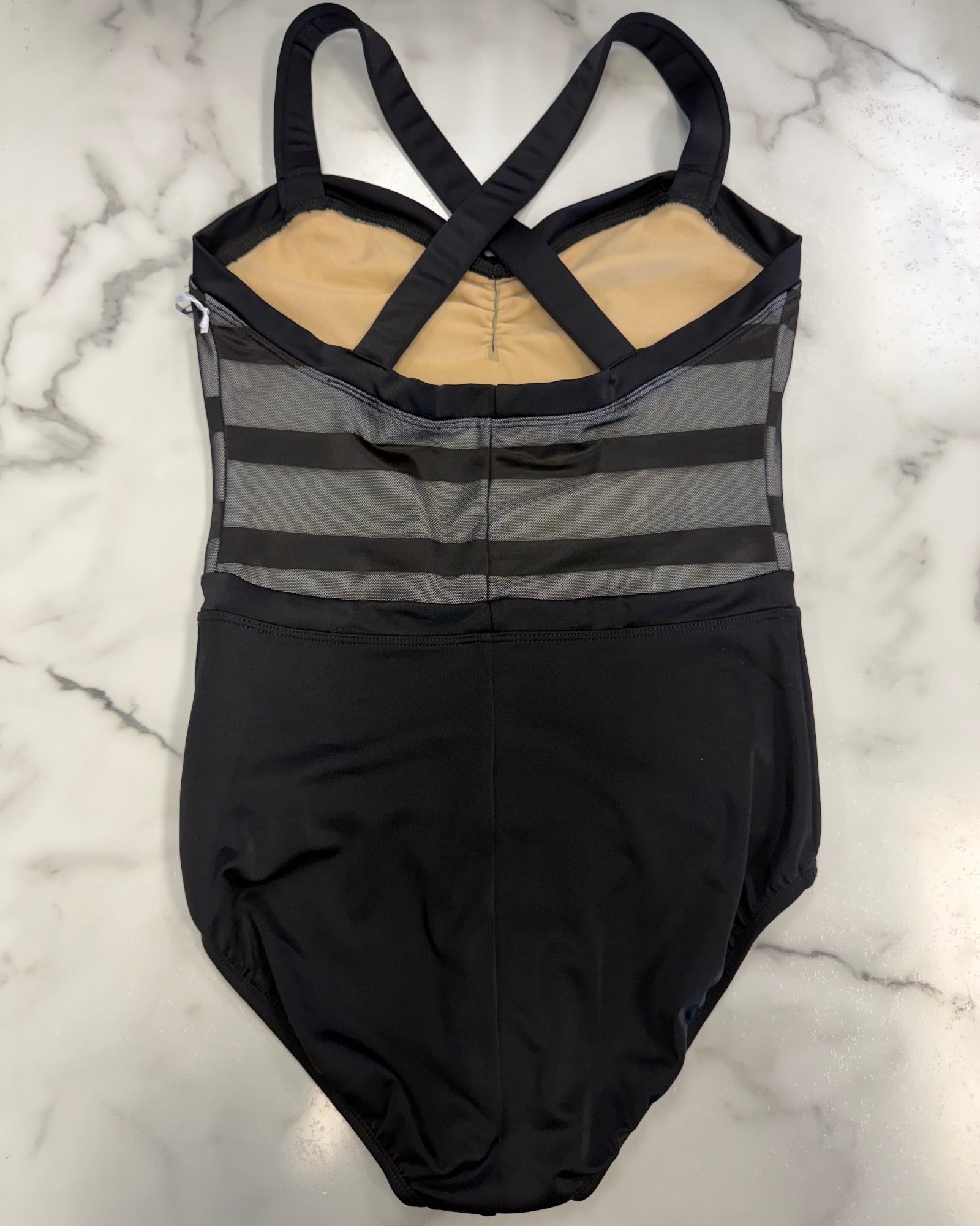 A black leotard with a sheer gray striped bodice, crossed shoulder straps, and a beige lining, laid flat on a white marble surface.