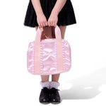A person wearing a black tutu skirt and black shoes holds a shiny light pink dance bag with pink handles against a plain white background.