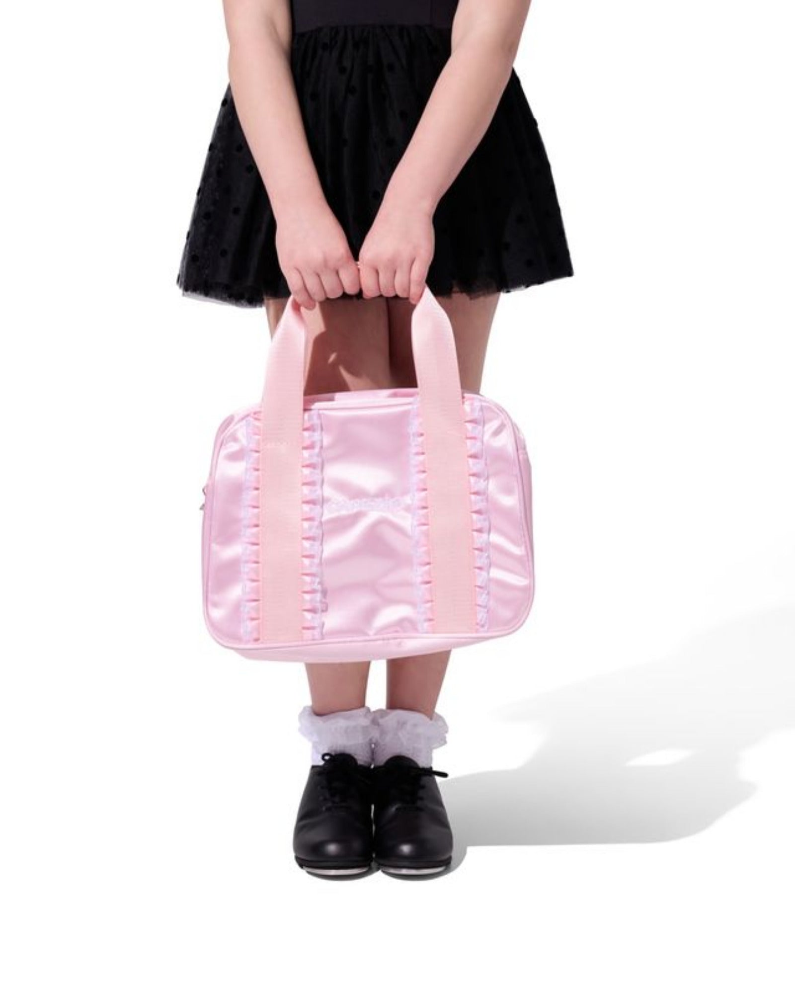 A person wearing a black tutu skirt and black shoes holds a shiny light pink dance bag with pink handles against a plain white background.