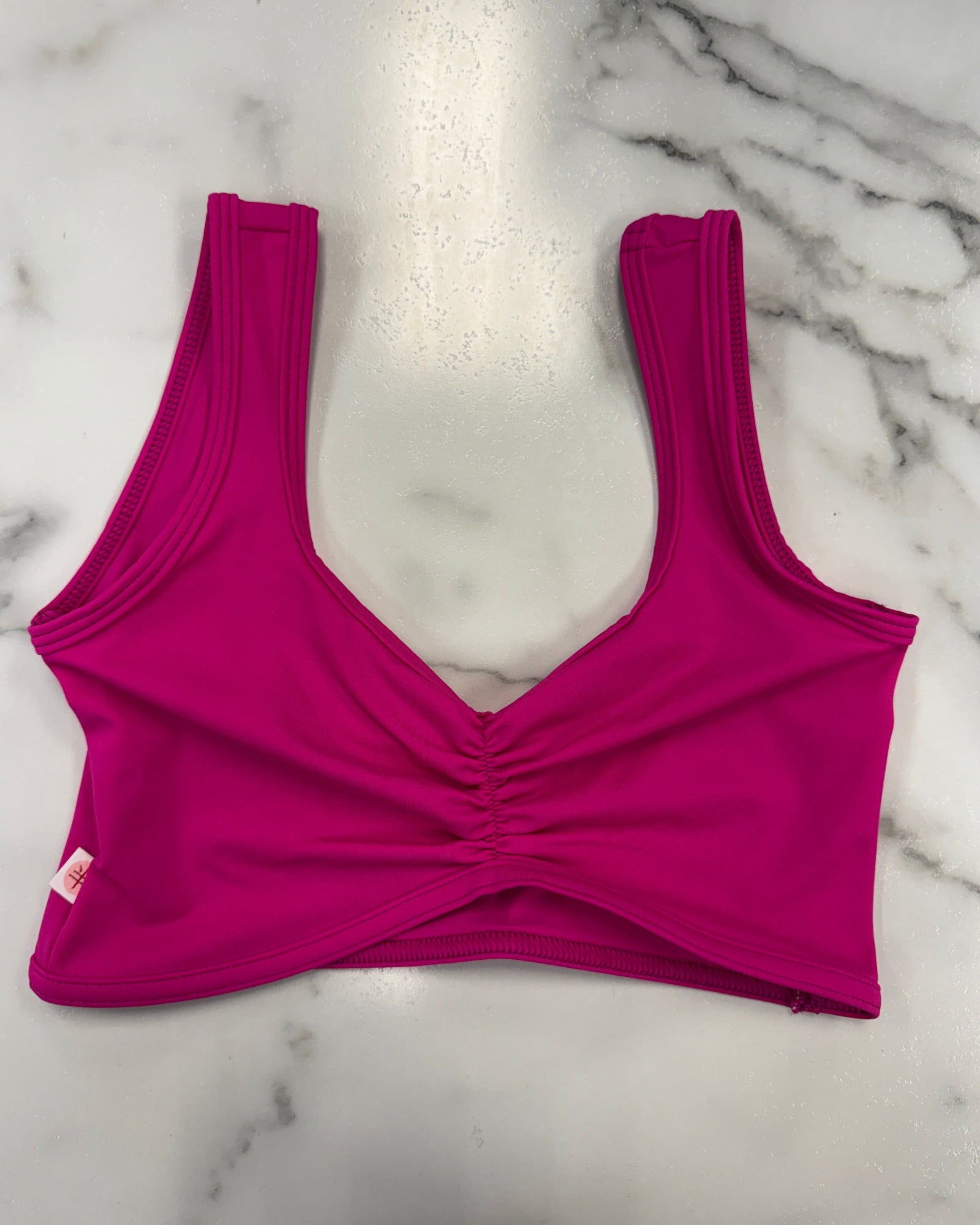 A bright magenta pink dance bra with a ruched front and wide shoulder straps, laid flat on a white marble surface.