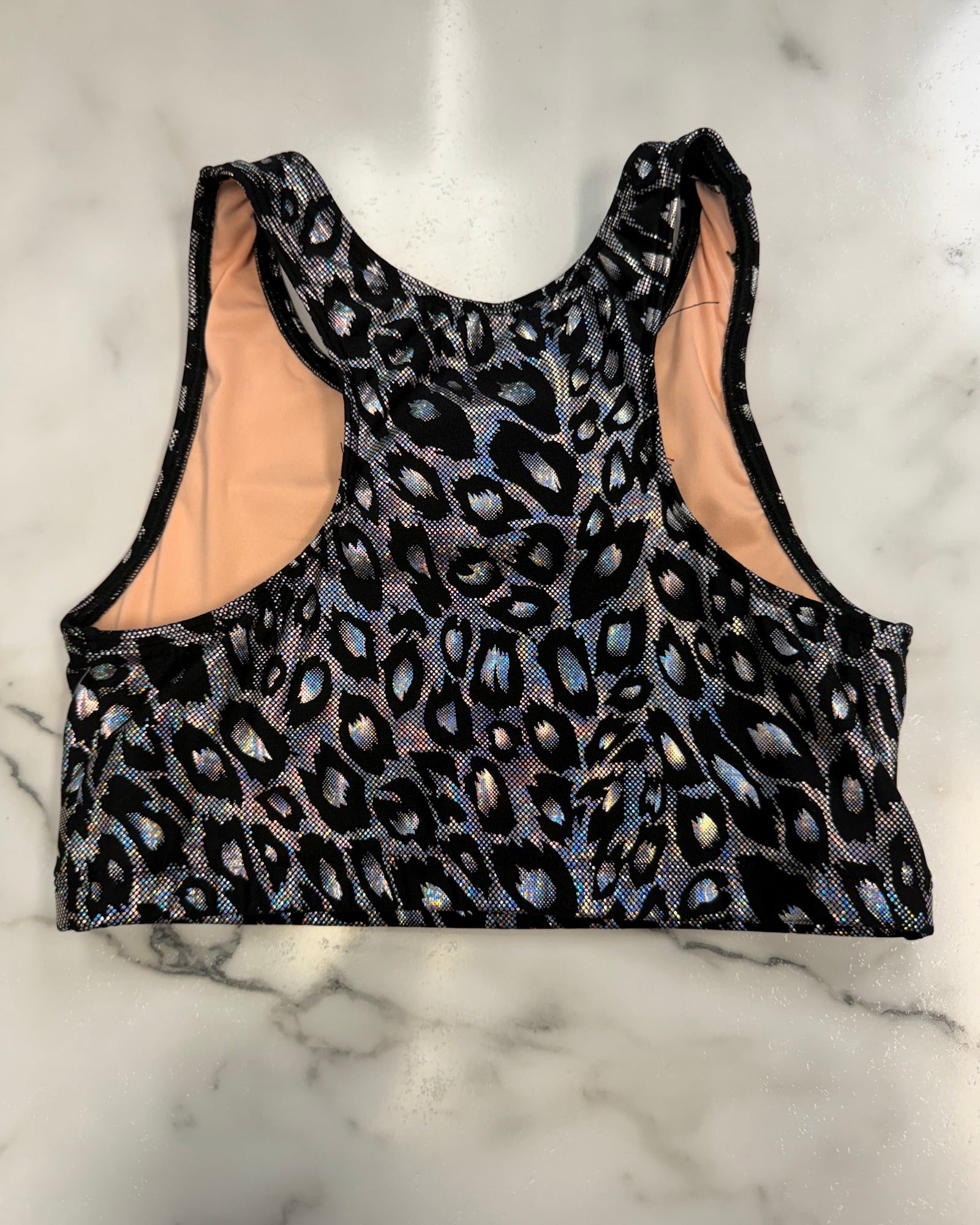 A sleeveless athletic crop top with a black leopard print pattern featuring iridescent blue and pink highlights, laid flat on a white marble surface.