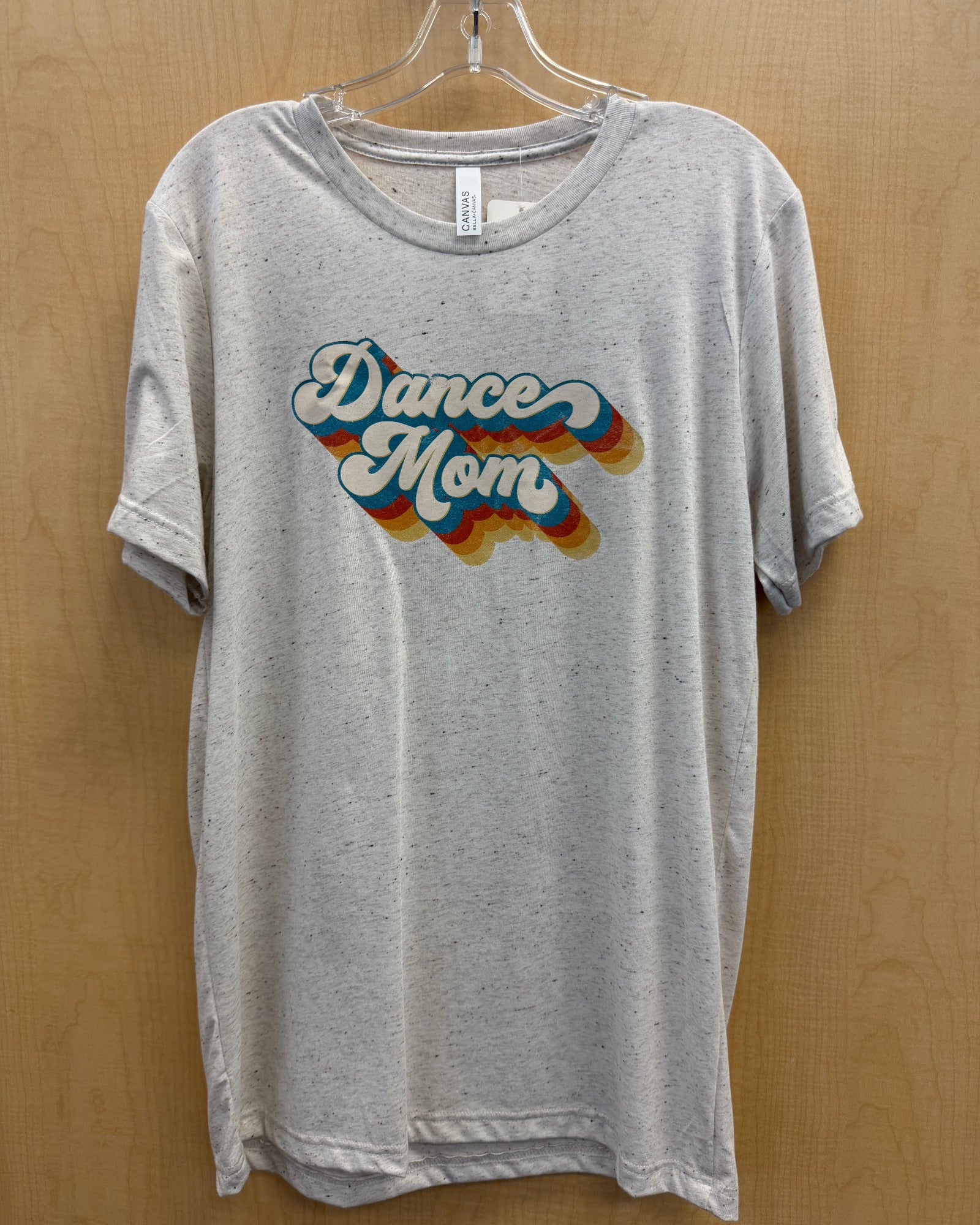 A light heather gray t-shirt hanging on a clear plastic hanger against a light wood-paneled wall. The shirt features a retro-style graphic on the chest that reads 'Dance Mom' in white bubble letters with a blue outline and a wavy rainbow shadow effect.
