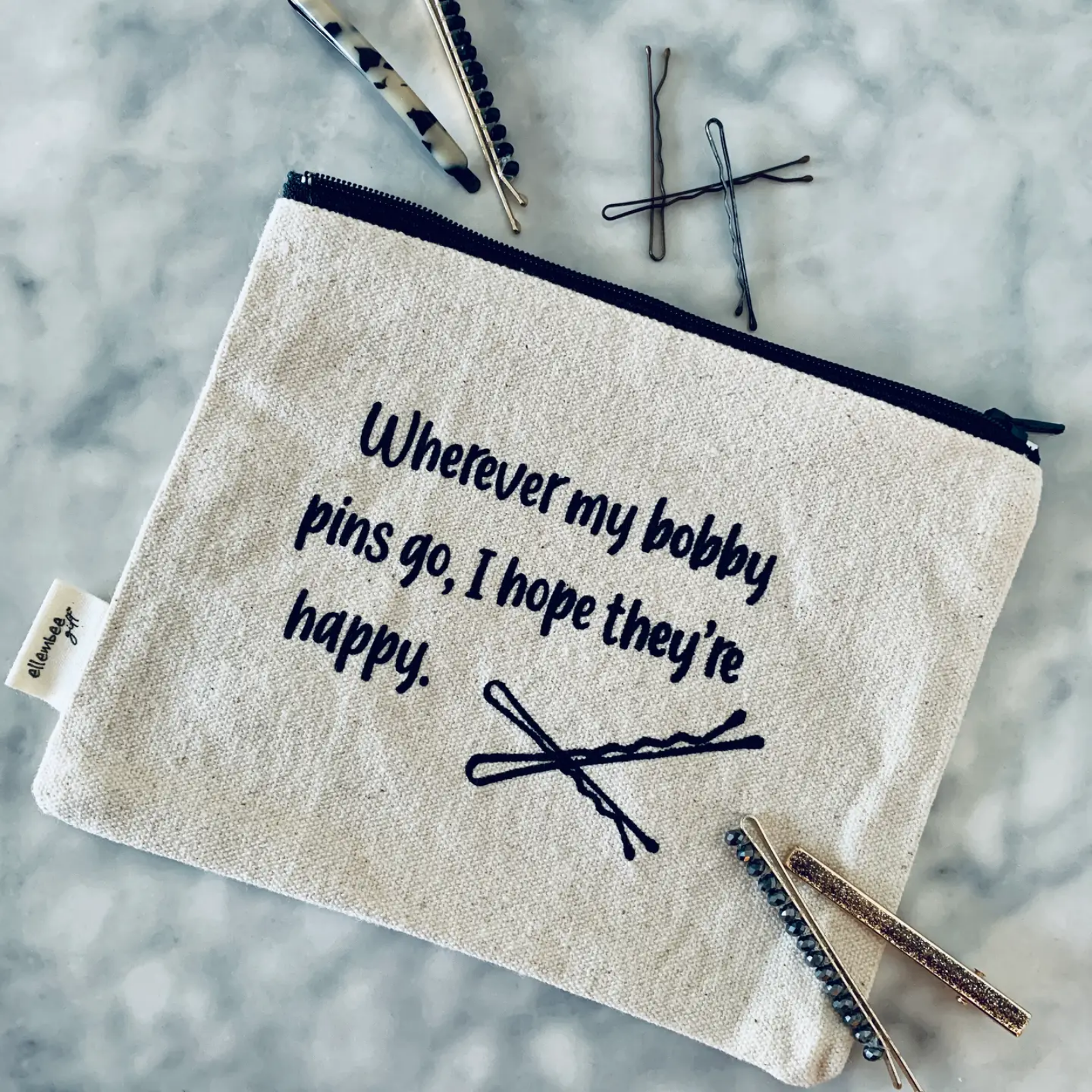 A canvas zippered pouch with a black zipper and embroidered text reading 'Wherever my bobby pins go, I hope they're happy.' alongside a small bobby pin illustration.