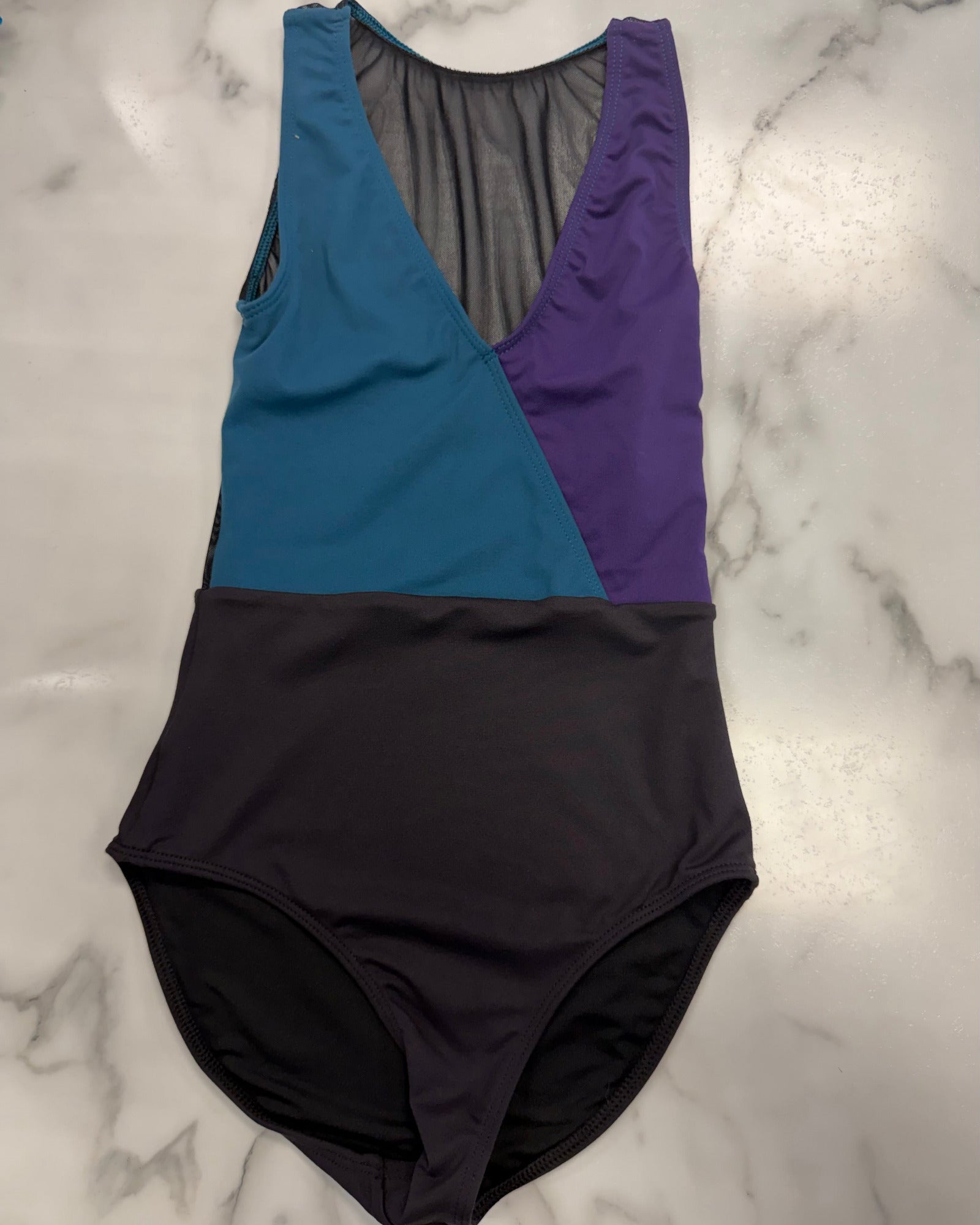 A color-blocked leotard laid flat on a white marble surface. The top portion features diagonal panels of teal blue, purple, and a sheer dark gray mesh insert, while the bottom is a solid dark charcoal black.