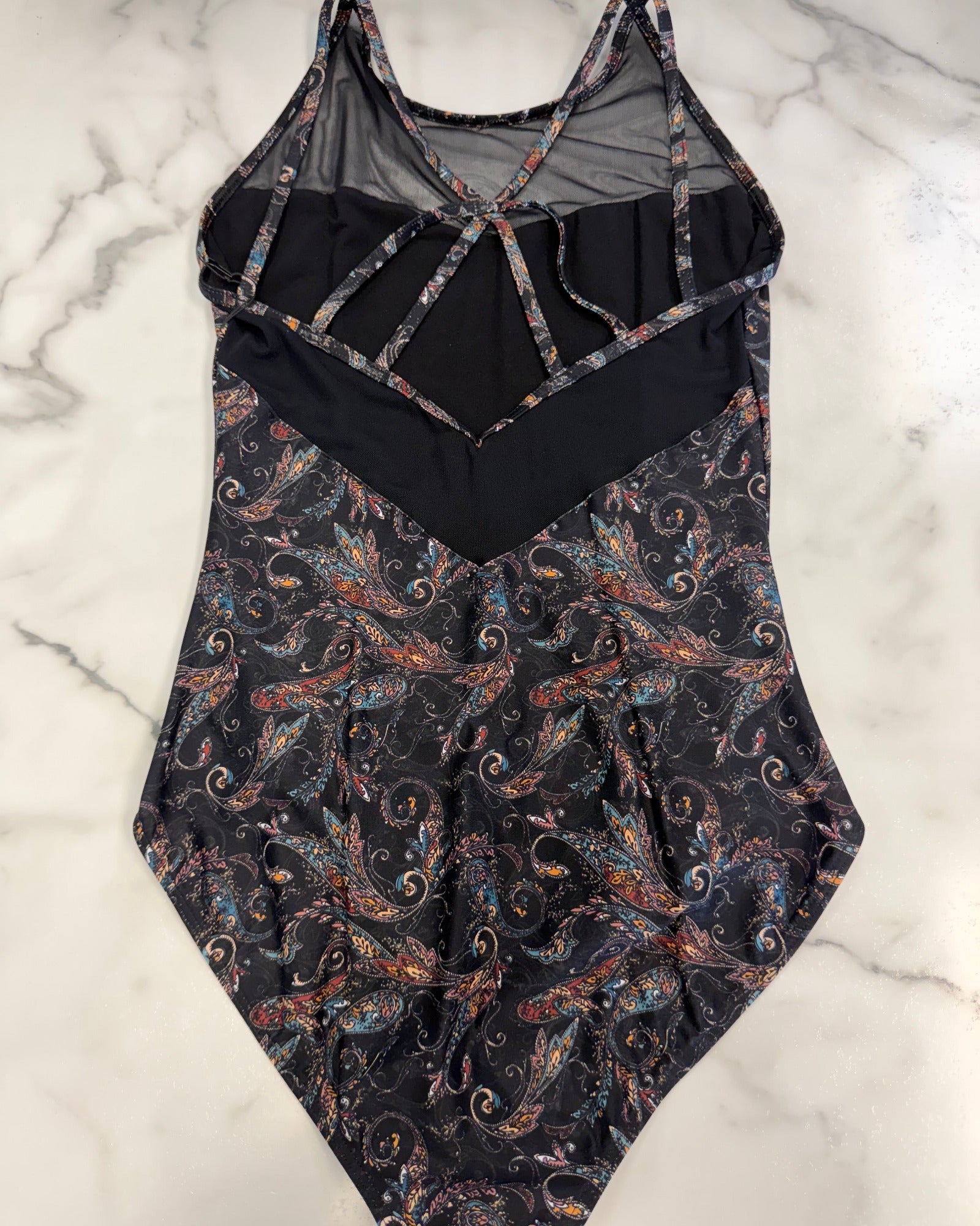 A leotard with a black mesh and solid black upper section, featuring a multicolor paisley pattern on a black base on the lower half, laid flat on a white marble surface.