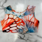 A sleeveless crop top with a vibrant butterfly wing pattern in orange, red, blue, and white, laid flat on a white marble surface.