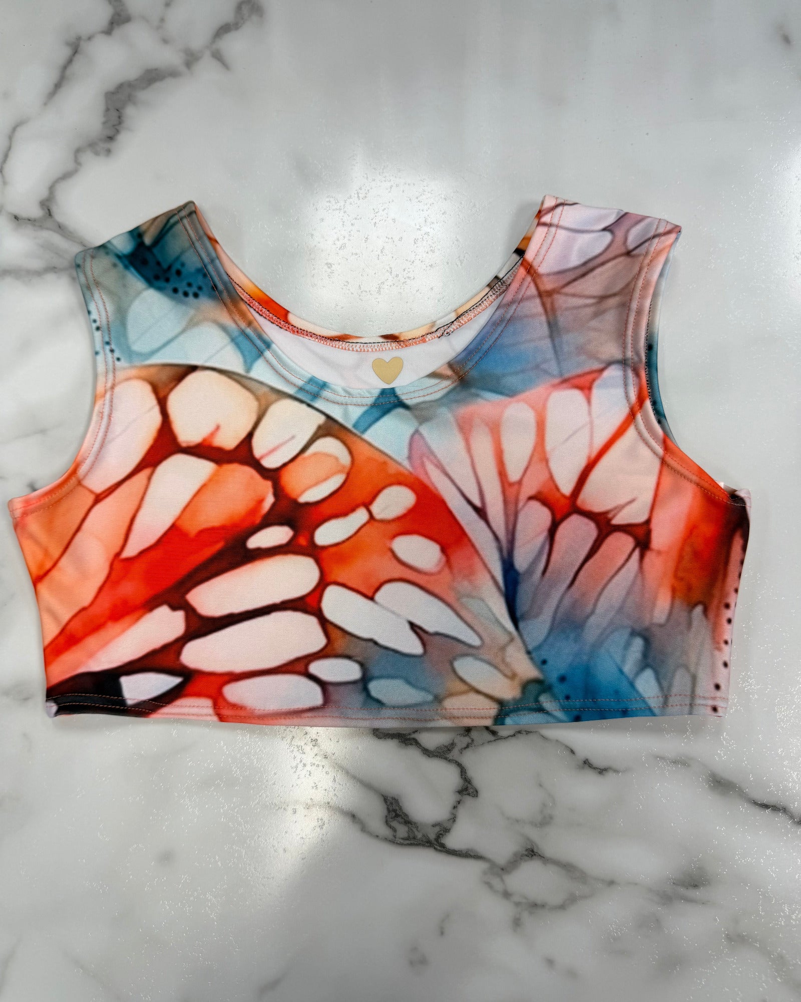 A sleeveless crop top with a vibrant butterfly wing pattern in orange, red, blue, and white, laid flat on a white marble surface.