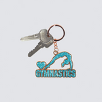 A gymnastics-themed keychain with a turquoise glitter emblem of a gymnast and the word 'GYMNASTICS' on a rose gold metal base, attached to a split key ring with two keys, shown on a light gray background.