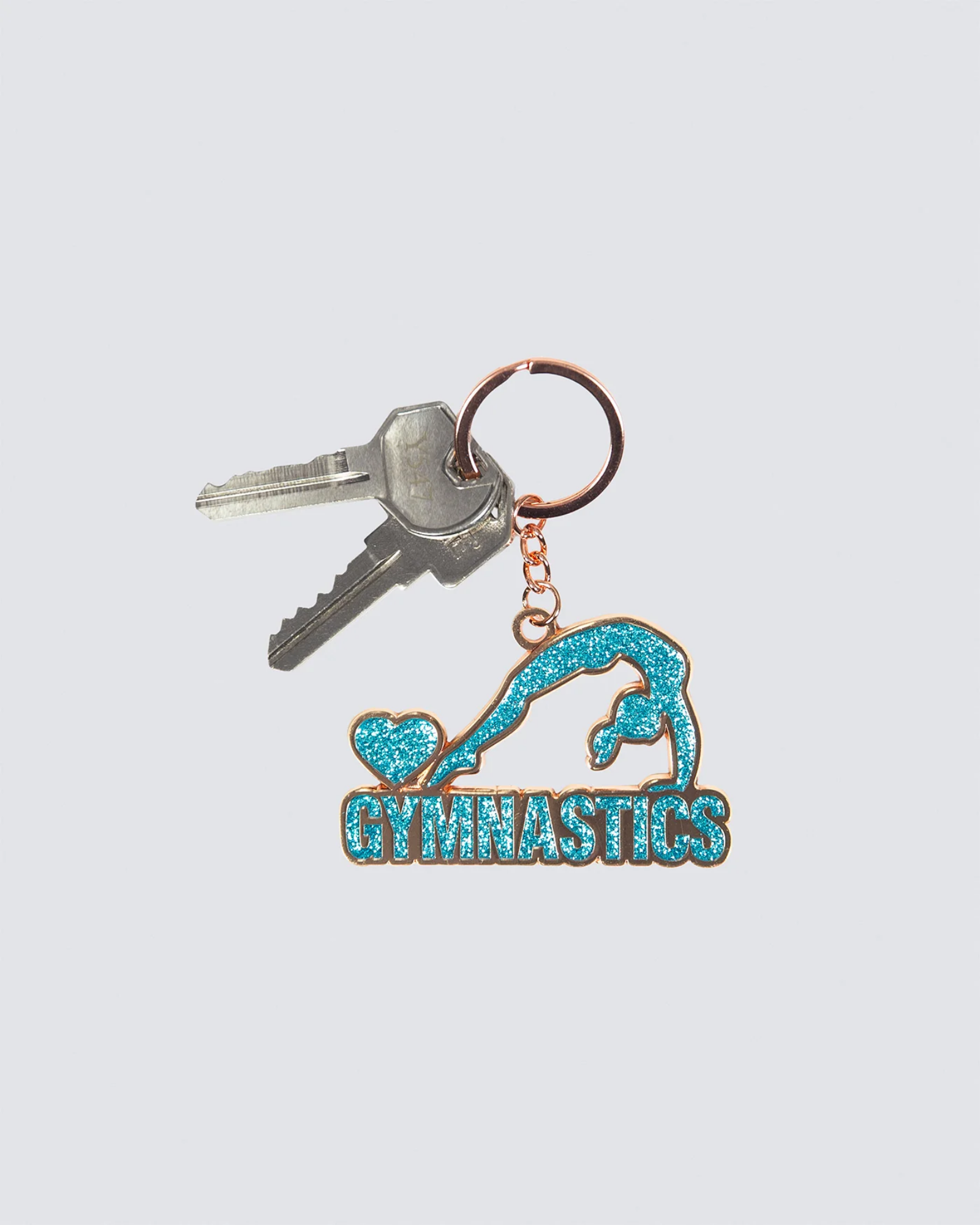 A gymnastics-themed keychain with a turquoise glitter emblem of a gymnast and the word 'GYMNASTICS' on a rose gold metal base, attached to a split key ring with two keys, shown on a light gray background.