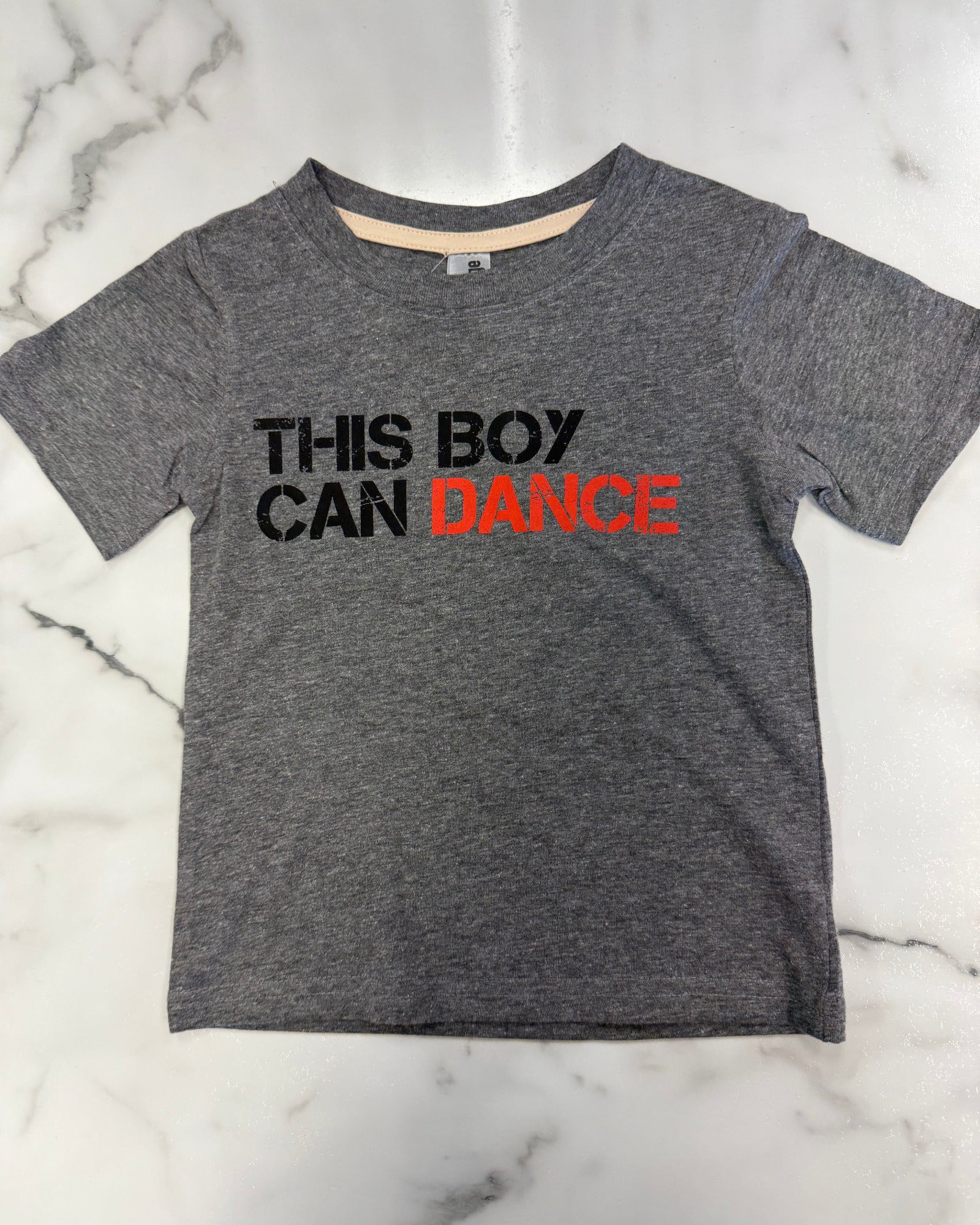 A toddler's heather gray short-sleeve t-shirt with the text 'THIS BOY CAN DANCE' printed on the chest, laid flat on a white marble background.