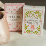 A pair of pale pink satin pointe shoes and two decorative boxes of dancing cards on a soft white surface. One box reads “PLAYING cards” and the other reads “dancer ROULETTE,” both with pink and green ballet-themed illustrations.