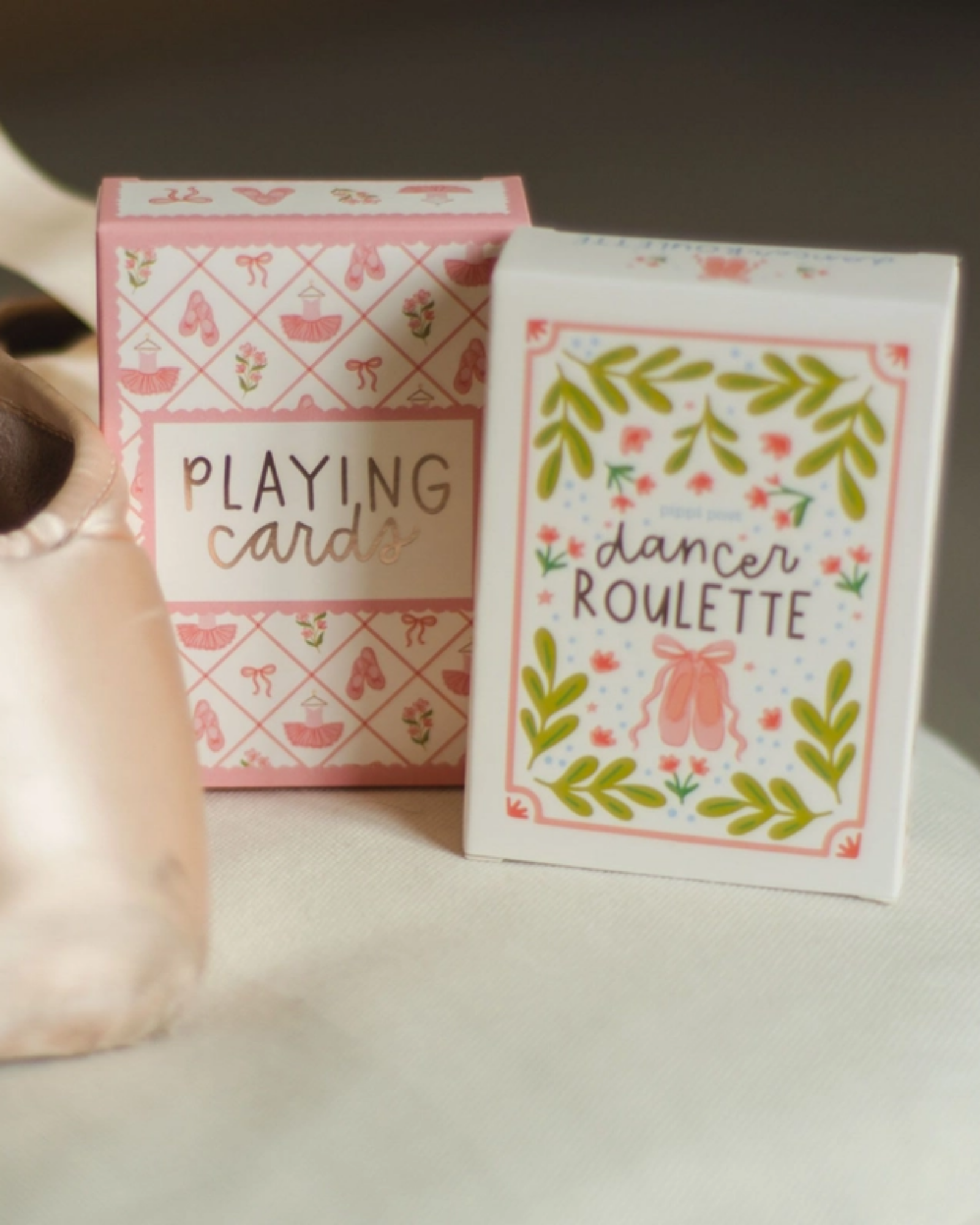 A pair of pale pink satin pointe shoes and two decorative boxes of dancing cards on a soft white surface. One box reads “PLAYING cards” and the other reads “dancer ROULETTE,” both with pink and green ballet-themed illustrations.