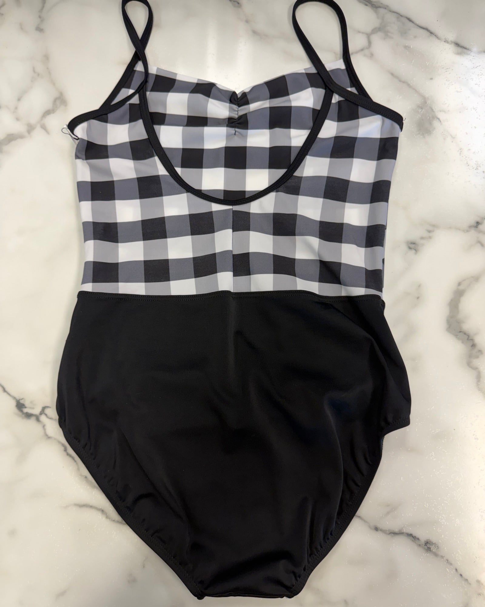 A leotard laid flat on a white marble surface. The top half features a black and white gingham check pattern with black straps, and the bottom half is solid black.