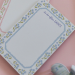A floral notepad with a blue border and the words 'to do after DANCE' printed in the top right, placed on a pink surface with other pastel notepads partially visible.