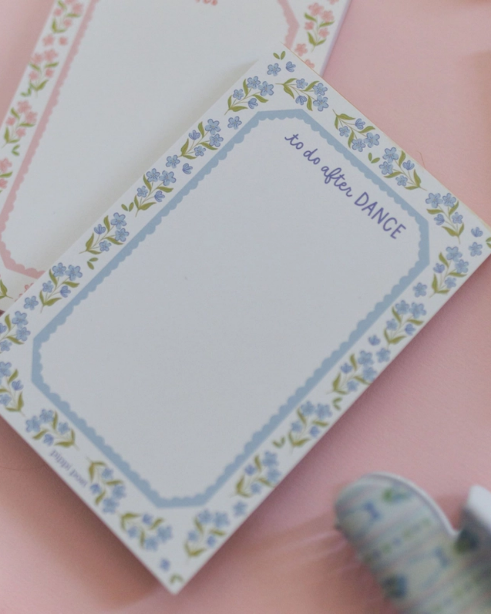 A floral notepad with a blue border and the words 'to do after DANCE' printed in the top right, placed on a pink surface with other pastel notepads partially visible.