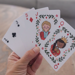 A hand holds four illustrated playing cards fanned out against a blurred indoor background. The cards are a 2 of clubs, 3 of clubs, 5 of diamonds, and a jack of diamonds, featuring cartoon characters and floral leaf borders.