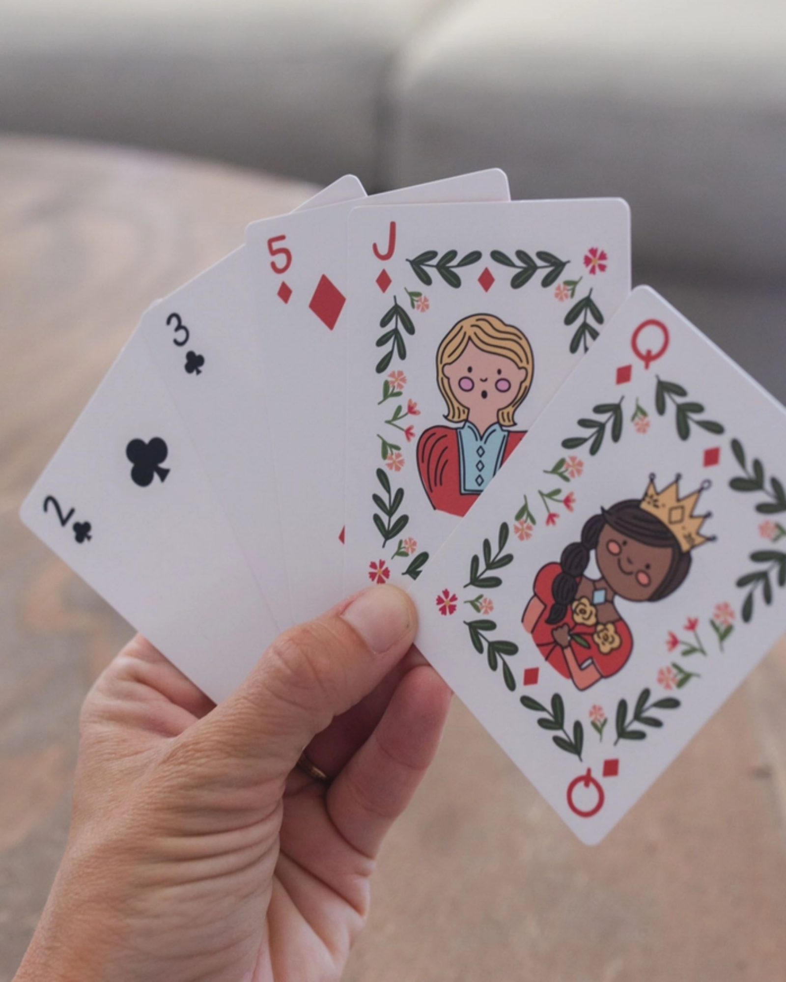 A hand holds four illustrated playing cards fanned out against a blurred indoor background. The cards are a 2 of clubs, 3 of clubs, 5 of diamonds, and a jack of diamonds, featuring cartoon characters and floral leaf borders.