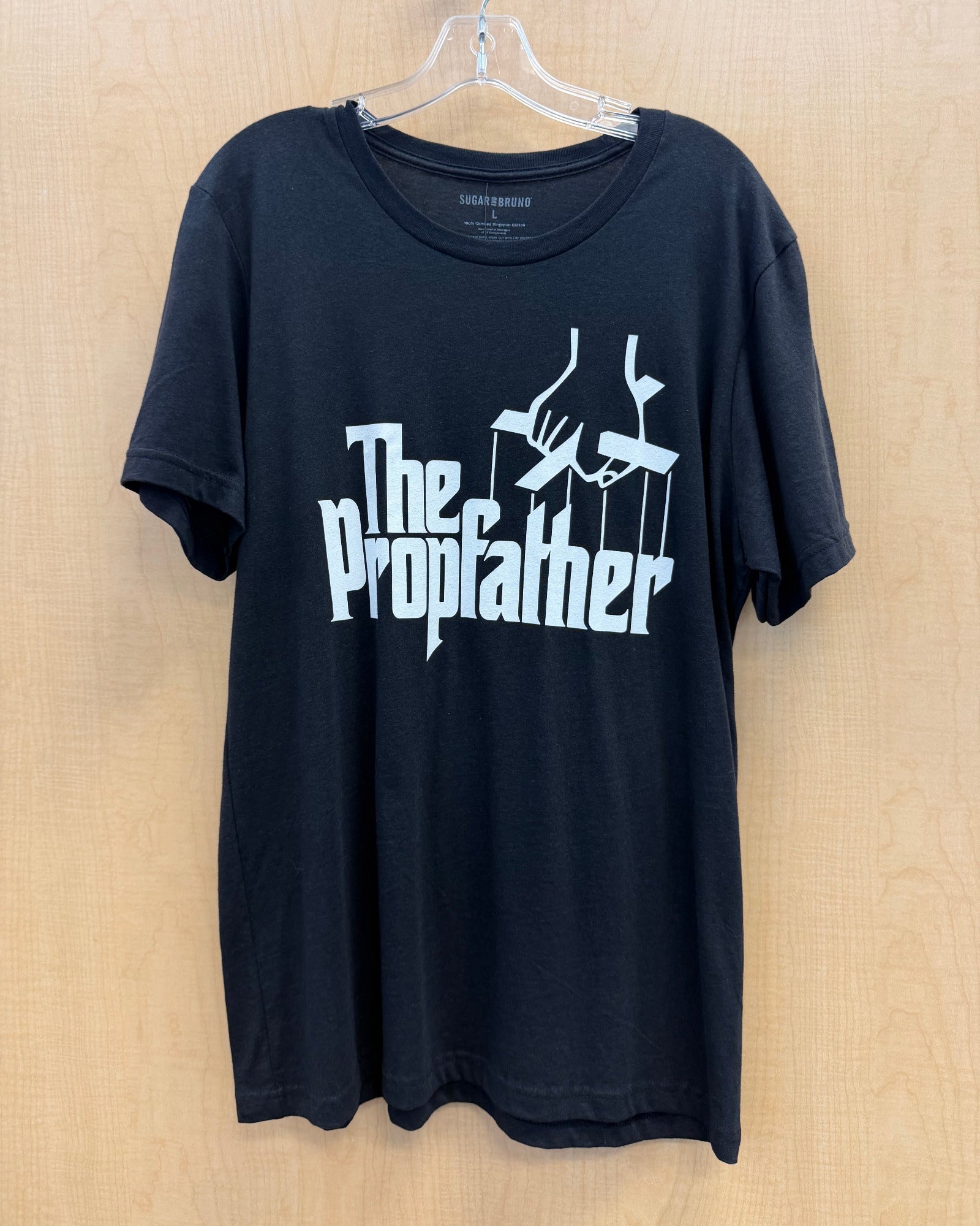 A black short-sleeve t-shirt hanging on a clear plastic hanger against a light wood-paneled wall. The shirt features a white graphic print on the front with the text 'The Propfather' and a stylized puppet hand design.