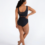 A woman modeling a black one-piece leotard from the back, standing in ballet slippers against a plain white studio background.