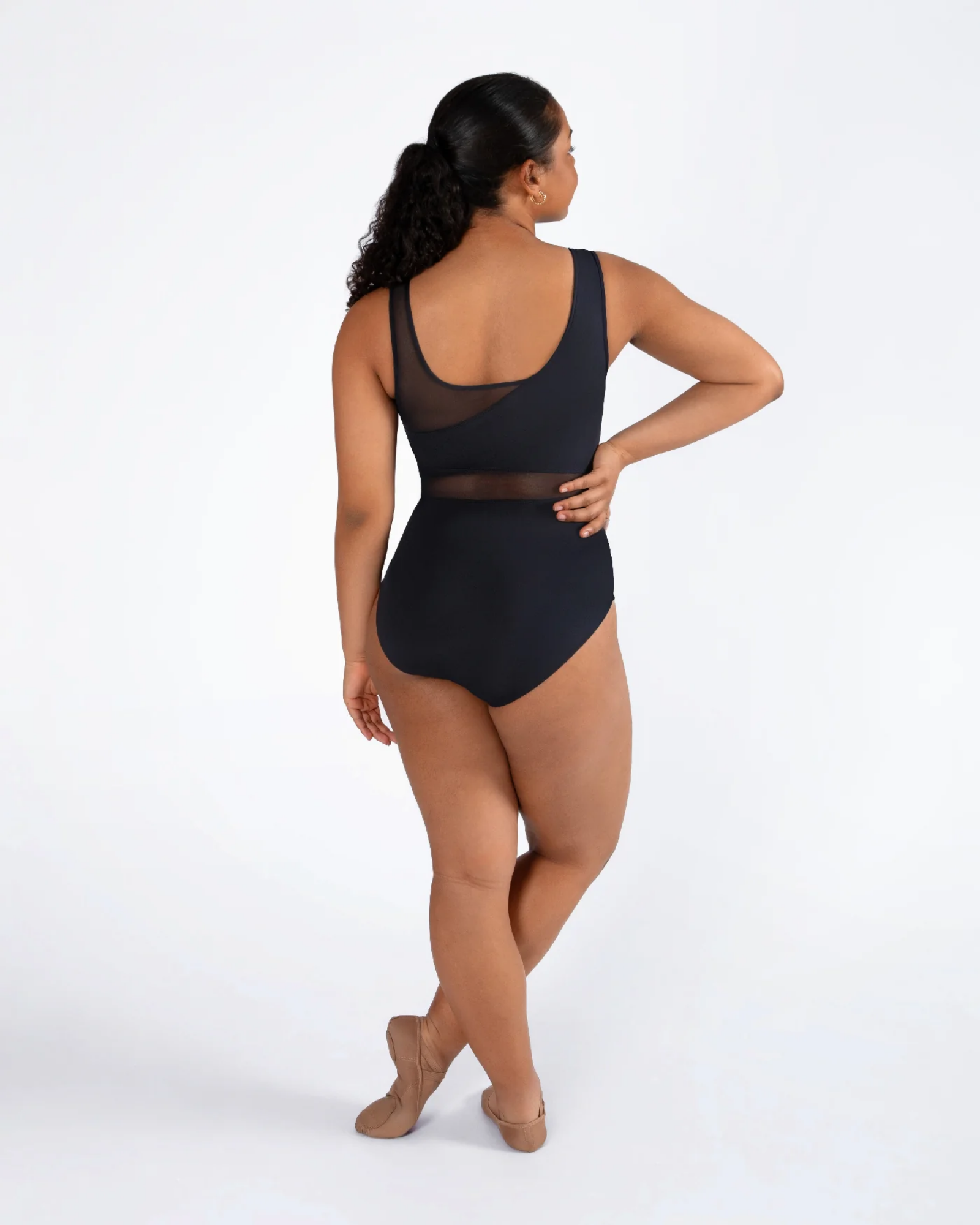 A woman modeling a black one-piece leotard from the back, standing in ballet slippers against a plain white studio background.