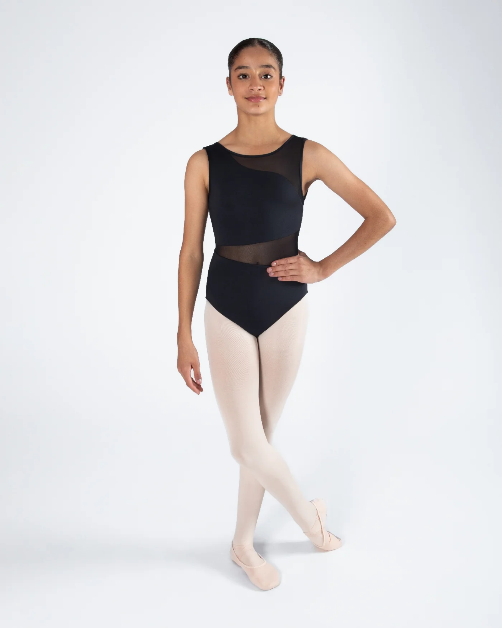 A young ballerina model wearing a black sleeveless leotard with sheer mesh panels and light pink tights, standing in a ballet position against a light gray studio background.