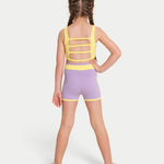 A young girl with braided hair is shown from the back wearing a two-piece dance set with a yellow strappy back and lavender-purple shorts with yellow trim, set against a plain light gray background.