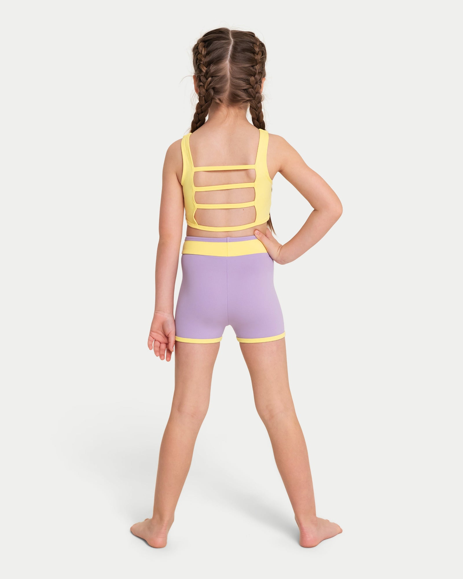 A young girl with braided hair is shown from the back wearing a two-piece dance set with a yellow strappy back and lavender-purple shorts with yellow trim, set against a plain light gray background.