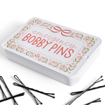 A white rectangular tin labeled 'emergency BOBBY PINS' with a pink bow and floral border, shown on a white background with several black bobby pins scattered in front.